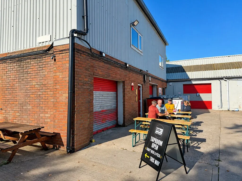 Benches and a Taproom Open A-board outside the New Invention brewery unit in Bloxwich, with people sitting at a table in the sun