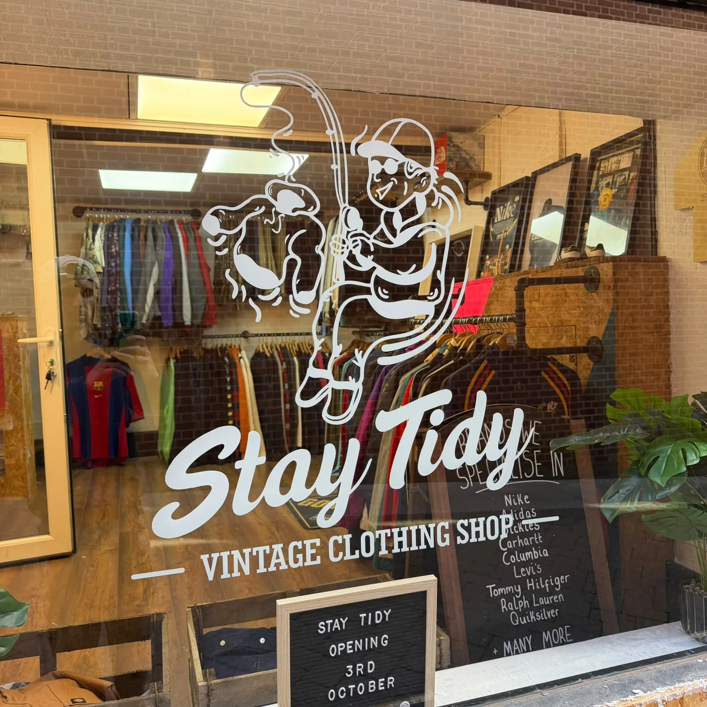 The Stay Tidy shop window, painted with a cartoon of an angler reeling in a jacket above the words Stay Tidy Vintage Clothing Shop, with rails of clothes and a Barcelona football shirt visible inside and a letter board reading Stay Tidy opening 3rd October