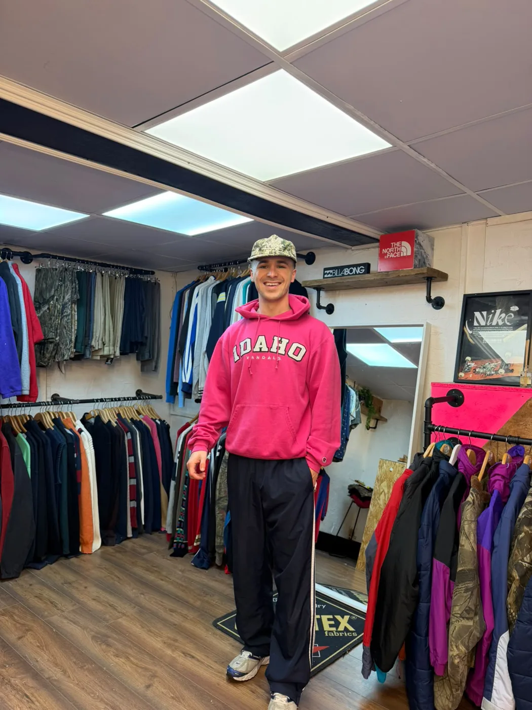 A man in a pink Idaho hooded sweatshirt and camouflage cap standing between rails of vintage jackets and sweatshirts inside Stay Tidy, under a North Face box and a framed Nike poster
