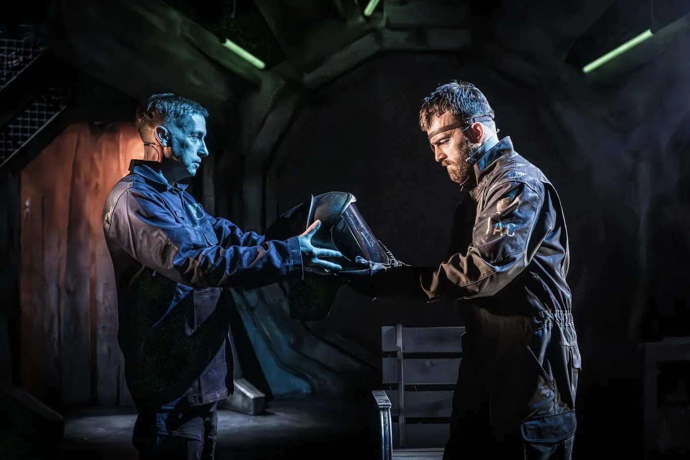 Two performers in worn flight suits on a dark stage set, one handing a helmet to the other