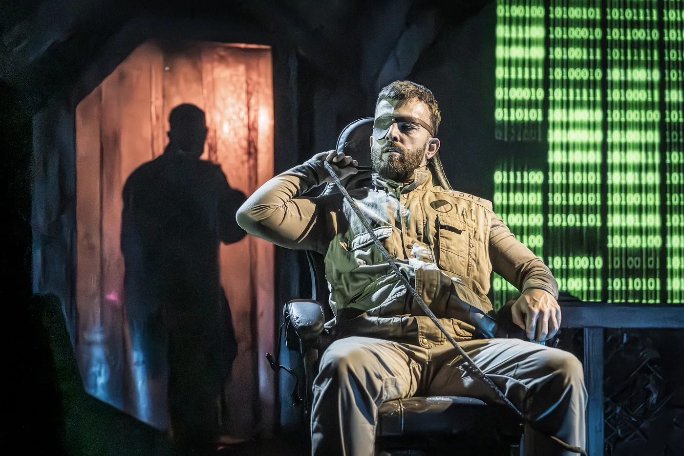 A performer seated in a chair on a dark stage, with a wall of green binary code projected behind him