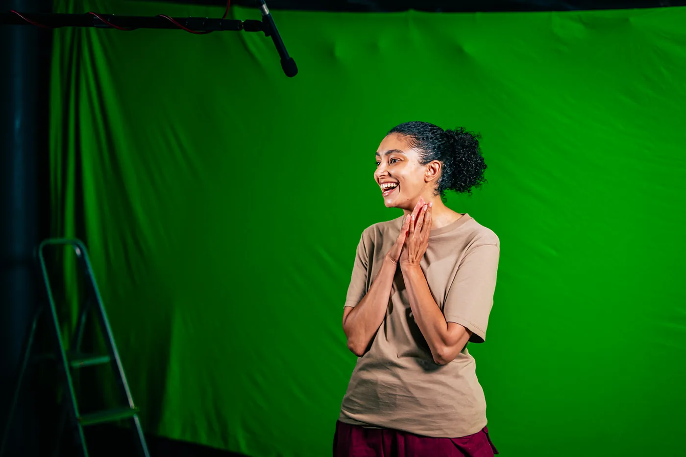 A performer standing in front of a lit green screen with hands together, a stepladder at the edge of the frame